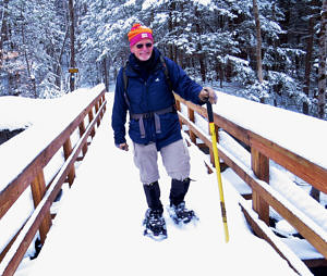 A smiling snowshoe hiker crosses a snowy footbridge. (photo © Meade Cadot)