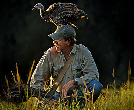 A man crouches with a Wild Turkey perched on his head. (photo © PBS Nature)