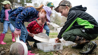 A group of children hold kitchen strainers and peer into plastic bins to look at aquatic wildlife gathered from a nearby pond. (photo © Ben Conant)