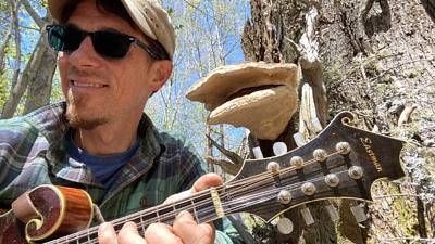 John Benjamin playing the mandolin near a mushroom.
