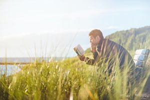 A man reading a book in a lakeside meadow. (photo © Ben White via unsplash)