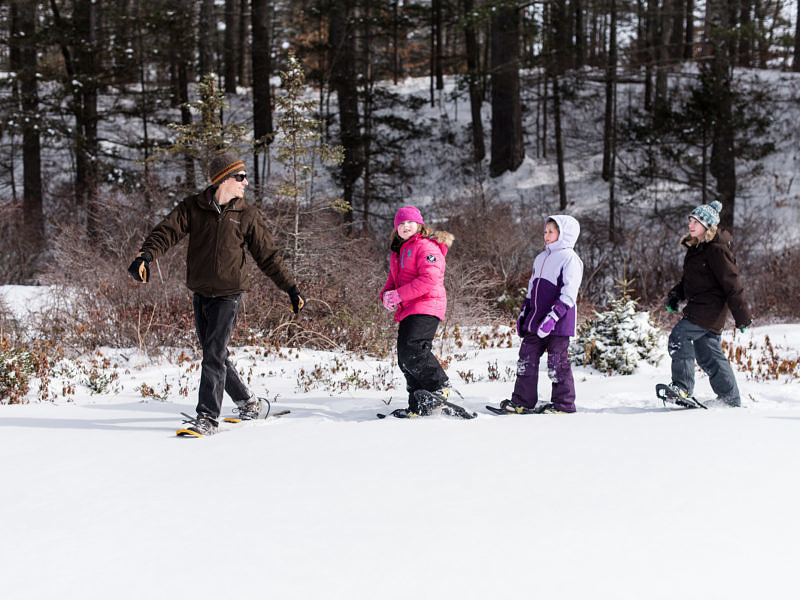A group of kids walks through a snowy field on snowshoes, with woods in the background. (photo © Ben Conant)