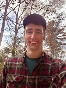 Joe Wiley, in a plaid flannel shirt and cap, standing in front of several trees.