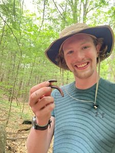A young man wearing a brimmed hat and striped shirt smiles while holding up a millipede.