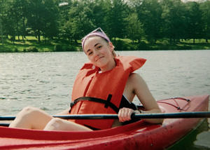 A young woman sits in a red kayak, holding a paddle and wearing an orange life vest.