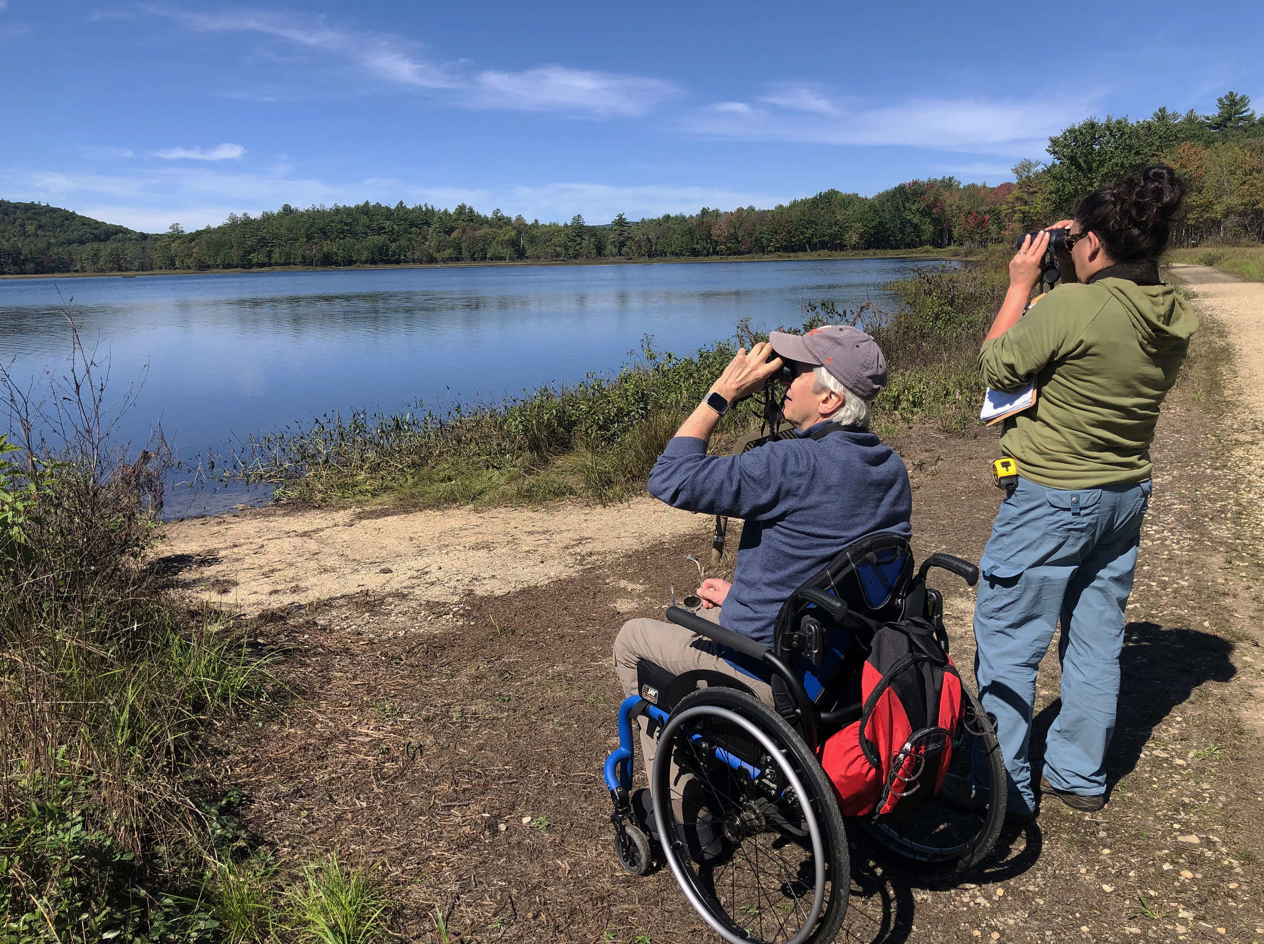 Two people -- one standing, one in a wheelchair -- hold binoculars up to their eyes while birding at a large lake. (photo © Jim Hassinger)