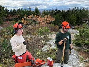 Two people in hard hats and chaps take a break from working a chainsaw during a view-clearing workday at the summit of Pack Monadnock. (photo © Phil Brown)