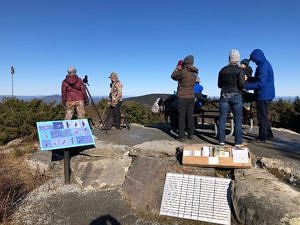 A group of people standing on a stone platform, holding binoculars to their faces and looking out into a blue sky. (photo © Phil Brown)