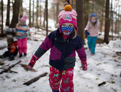 A child wearing a face mask and winter clothing walks through snowy woods. (photo © Ben Conant)