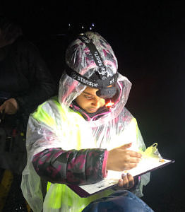 A child wearing a headlamp, rain poncho, and reflective vest looks down at a clipboard. (photo © Karen Seaver)