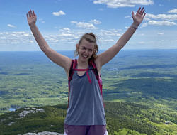Rachel Ranelli, standing on the summit of Mount Monadnock with her arms in the air on a bluebird day.