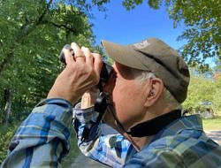 A side view of a man looking through binoculars, with a hearing aid visible in his left ear. (photo © Sandy Taylor)