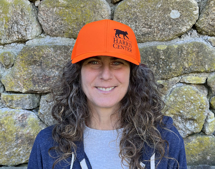 Susie Spikol standing in front of a stone wall, wearing a Harris Center trucker cap in blaze orange.