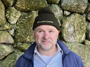 Eric Masterson, standing in front of a stone wall, wearing an olive-green wool hat with the Harris Center logo on it.