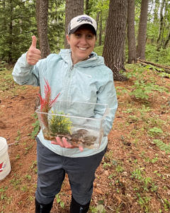 Jenn Sutton, standing in the woods. She is holding a plastic tank of turtles in her left hand, and giving a "thumb's up" sign in her right hand. 