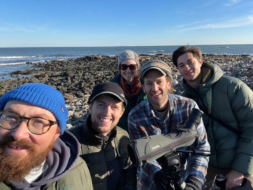 Five birders smile at the camera with a scope in the foreground and the coast behind them (photo: Chad Witko)
