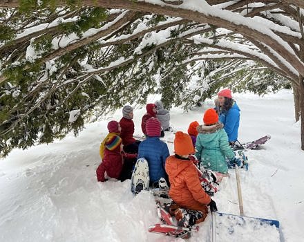 Several children sit with Susie Spikol under the eaves of a snowy yew tree (photo © Karen Rent).