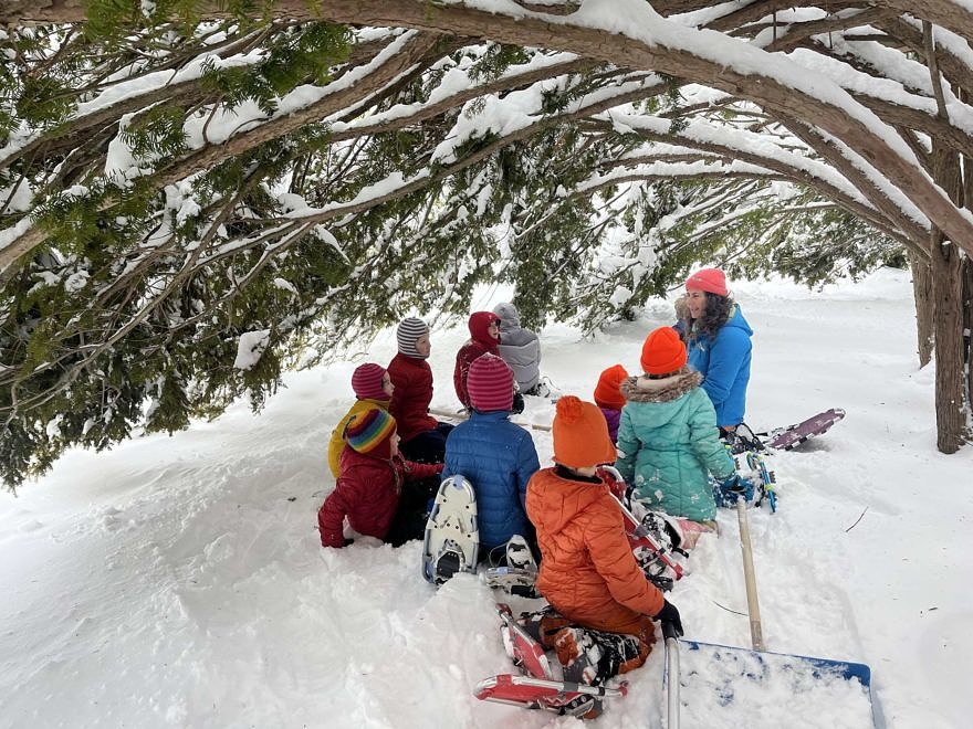 Several children sit with Susie Spikol under the eaves of a snowy yew tree (photo © Karen Rent).