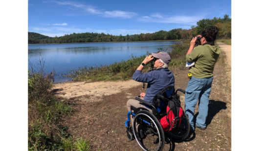Two people -- one standing, one in a wheelchair -- hold binoculars up to their eyes while birding at a large lake. (photo © Jim Hassinger)