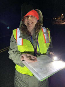 Karen Seaver stands on a dark road, wearing a reflective vest. In one hand, she is holding a clipboard with a data form. In the other hand, a spring peeper. (photo © Taylor Jackson)