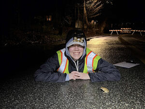 A person in a reflective vest lies on the pavement next to an American toad, with road barricades visible in the background. (photo © Brett Amy Thelen)