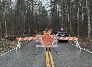 A person wearing a reflective vests stands in a road behind a barricade and a sign that says "Road Closed." (photo © Brett Amy Thelen)