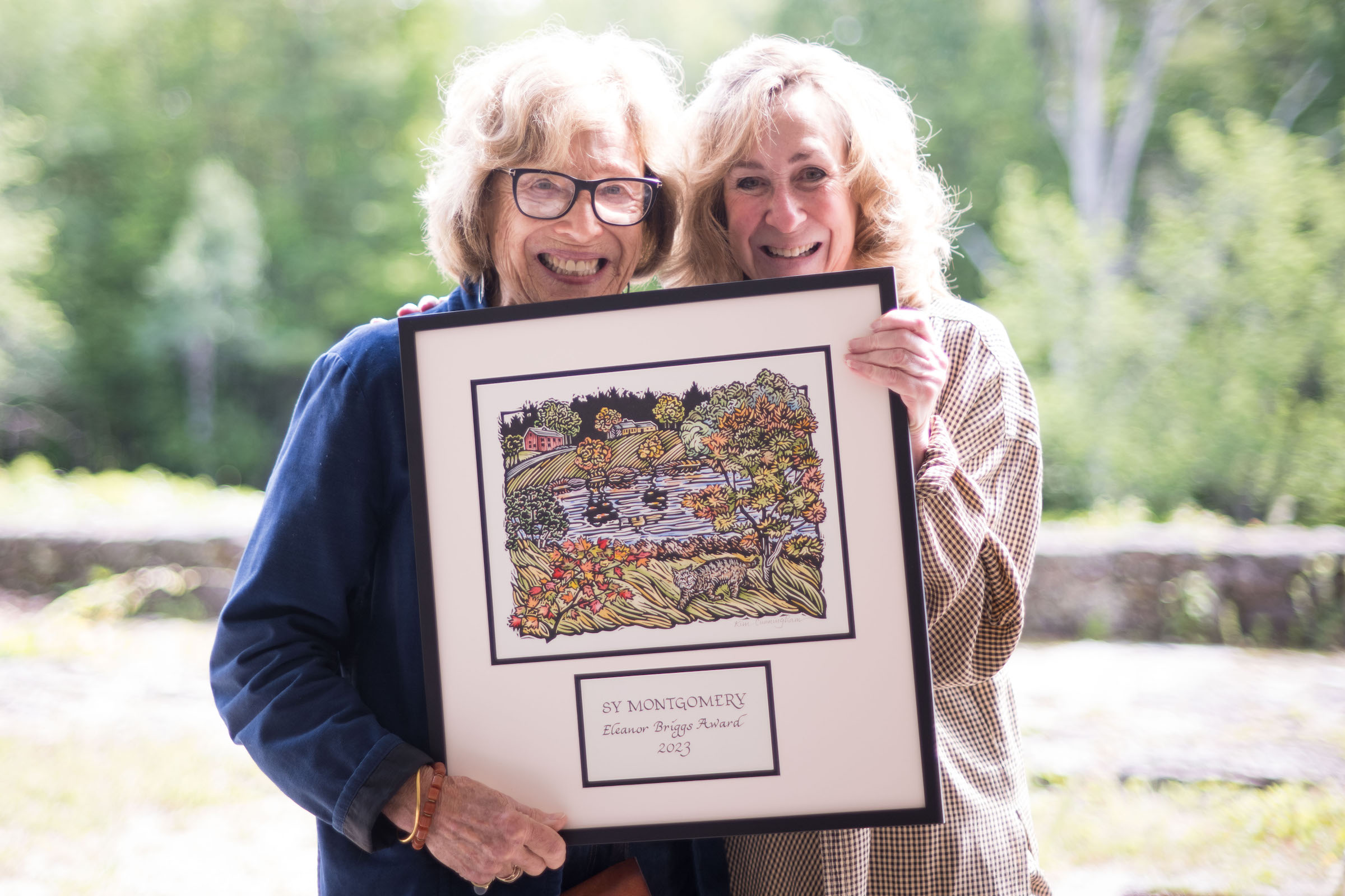 Two smiling women, Eleanor Briggs and Sy Montgomery, hold the Eleanor Briggs Award, a Lino print plaque, matted and framed. (photo: Ben Conant)