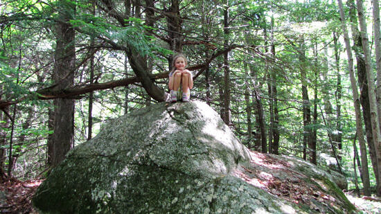 A child smiles atop a large boulder in the forest. (photo: Jenna Spear)