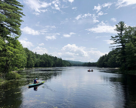 A kayaker and two people in a canoe paddle a waterbody surrounded by forest. under a blue, cloud-filled sky. (photo: Brett Amy Thelen)