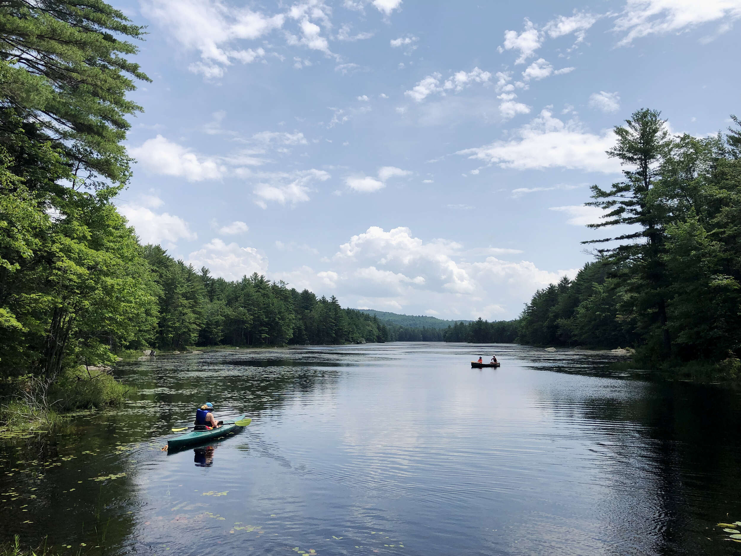 A kayaker and two people in a canoe paddle a waterbody surrounded by forest. under a blue, cloud-filled sky. (photo: Brett Amy Thelen)