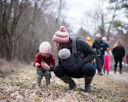 A mom bends down to show her toddler a small object, with a baby strapped to her front. (photo © Ben Conant)