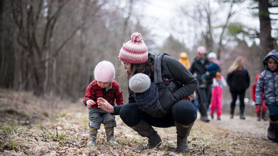 A mom bends down to show her toddler a small object, with a baby strapped to her front. (photo © Ben Conant)