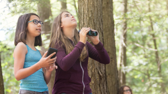 Two teenaged girls use binoculars in the forest. (photo © CanvaCommons)
