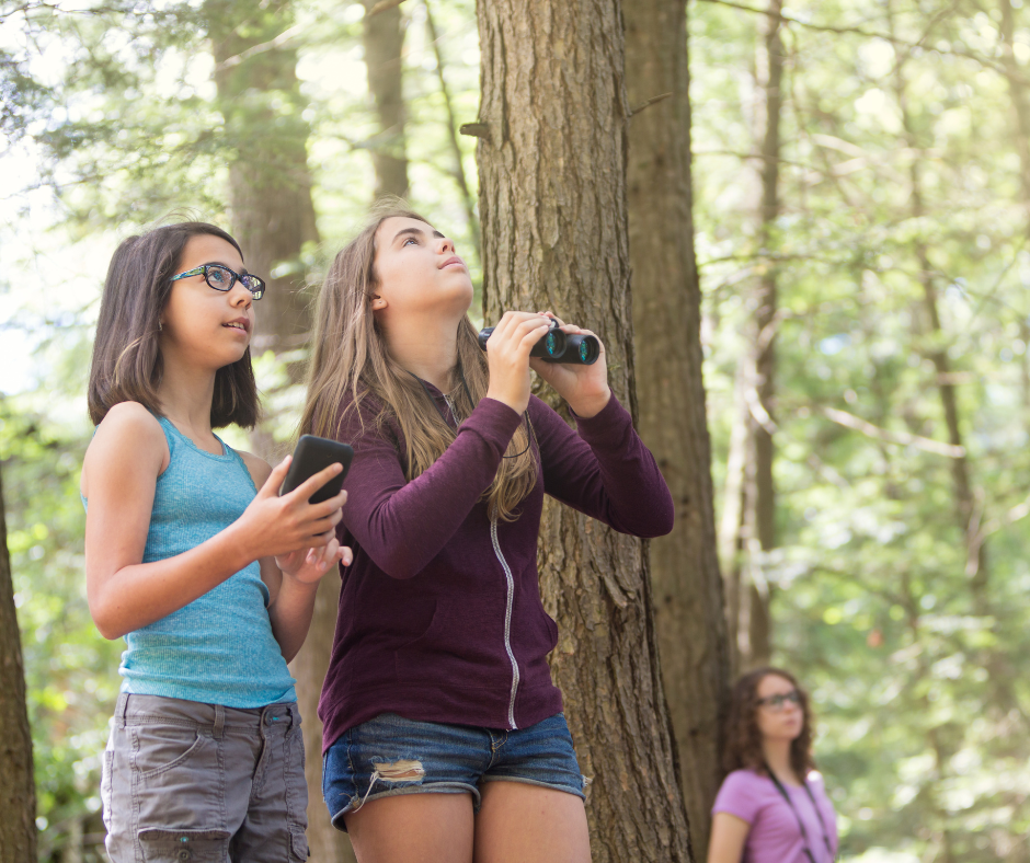Two teenaged girls use binoculars in the forest. (photo © CanvaCommons)