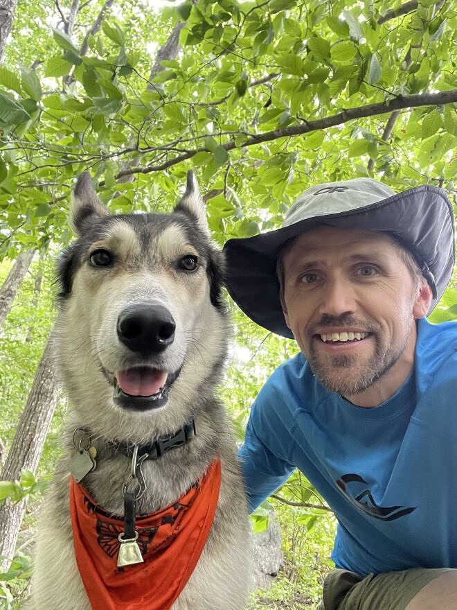 A wolf-like dog wearing and orange bandana, next to a smiling Miles. (photo © Miles Stahmann)