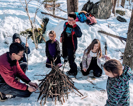 John Benjamin builds a fire in the snowy outdoors, with children watching. (photo © ?)