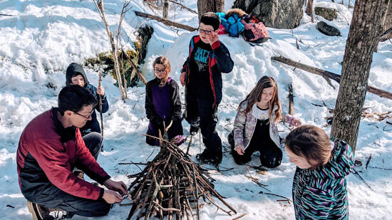John Benjamin builds a fire in the snowy outdoors, with children watching. (photo © ?)