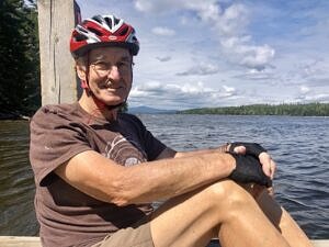 John Odgers, wearing a biking helmet and gloves, smiles in front of a waterbody. (photo © John Odgers)
