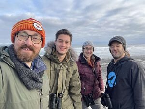 Four people dressed for winter smile at the camera, with a cloudy sky and the coast at their back. (photo © Chad Witko)
