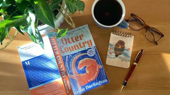 The book "Otter Country" lying on a table next to a coffee cup, a pair of glasses, a notebook, pen, and plant. (photo © Tin House)