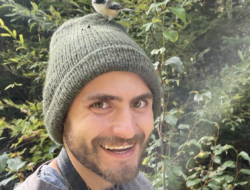 Chris Liazos, with a chickadee on his knit cap, smiles at the camera.
