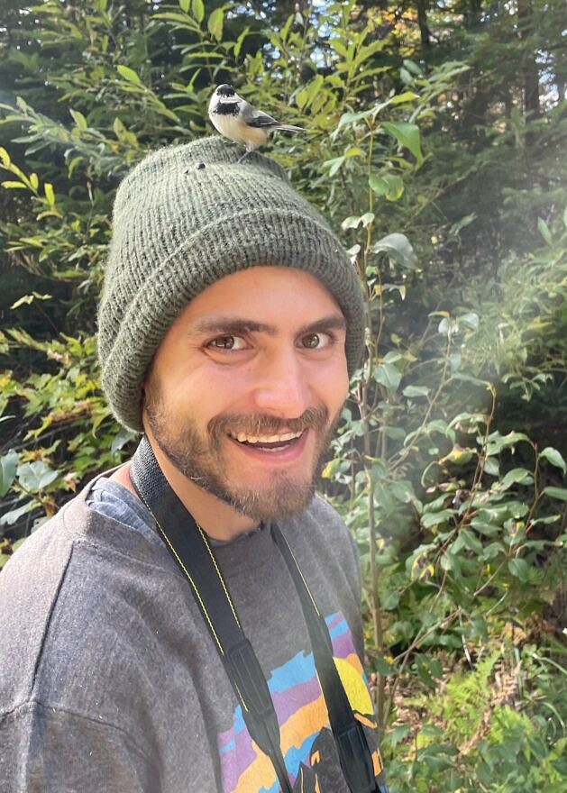 Chris Liazos, with a chickadee on his knit cap, smiles at the camera.