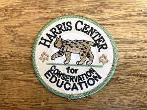 An embroidered patch with a bobcat and the words, "Harris Center for Conservation Education" against a wood grain background.