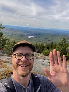 Sam Faller smiles and waves to the camera atop a mountain. 