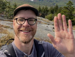 Sam Faller smiles and waves to the camera atop a mountain.