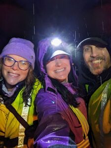 Three people smile while wearing reflective vests and headlamps, and standing outside on a rainy night. (photo © Jen Boisvert)