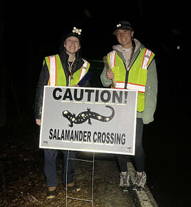 Two young women in reflective vests stand behind a sign that says, "Caution! Salamander Crossing" (photo © Brett Amy Thelen)