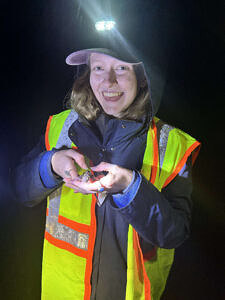 A young woman wearing a reflective vest and handlamp smiles while holding a spotted salamander in her hands. (photo © Chloe March)