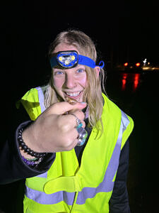 A young woman wearing a reflective vest smiles at the spring peeper she is holding in her hand. (photo © Chloe March)
