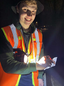 A young man wearing a reflective vest smiles while recording data on a small data sheet. (photo © TaylorJackson)